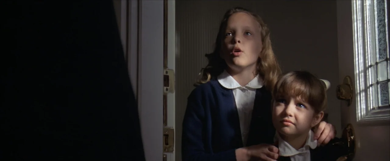 Arlington Road (1999) movie still: medium — A dark indoor entryway features an open door with a decorative glass panel. Two young girls stand in the…