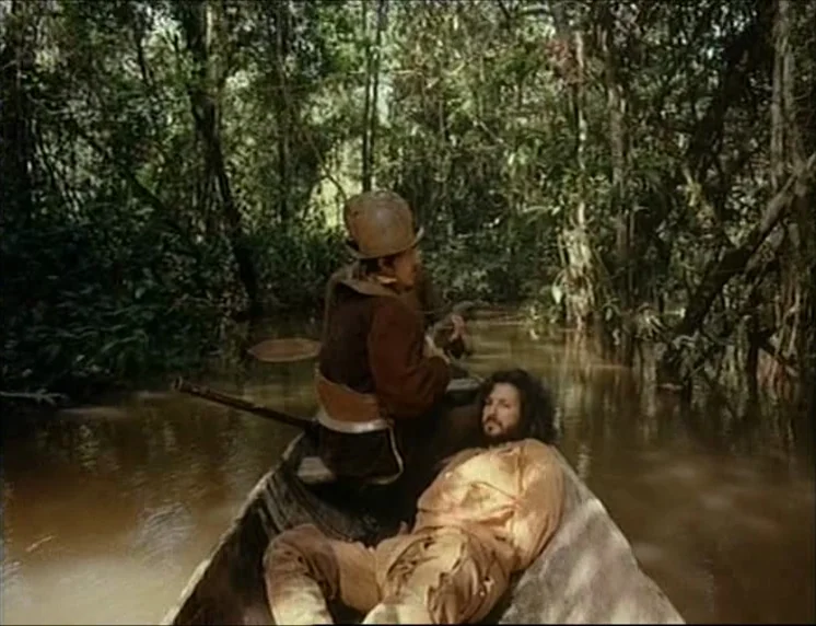 Aguirre, The Wrath Of God (Aguirre, der Zorn Gottes) (1972) movie still: fullbody — A wooden boat floats on a murky, brown river through a dense…