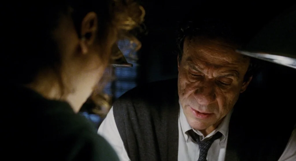 Mimic (1997) movie still: medium — A dark office illuminated by a single warm desk lamp at night. An older man in a sweater vest looks downward…