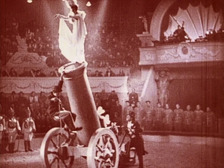 Innocence Unprotected (1968) movie still: wide — An indoor performance space with a large central cannon and audience-filled tiered seating. A…