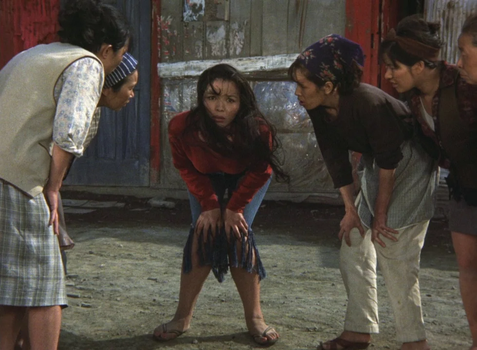 Dodes’ka-den (1970) movie still: medium — An outdoor alleyway with weathered walls serves as the backdrop for a gathering. A woman in a red shirt…