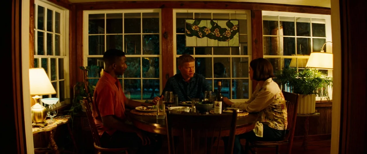 Kinds of Kindness (2024) movie still: medium — A wood-paneled room features a circular table where three people are dining at night. Multi-paned…