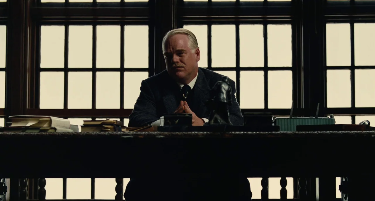 The Master (2012) movie still: medium — A man sits behind a heavy wooden desk in front of a large window. A sculpture and books are placed on the…