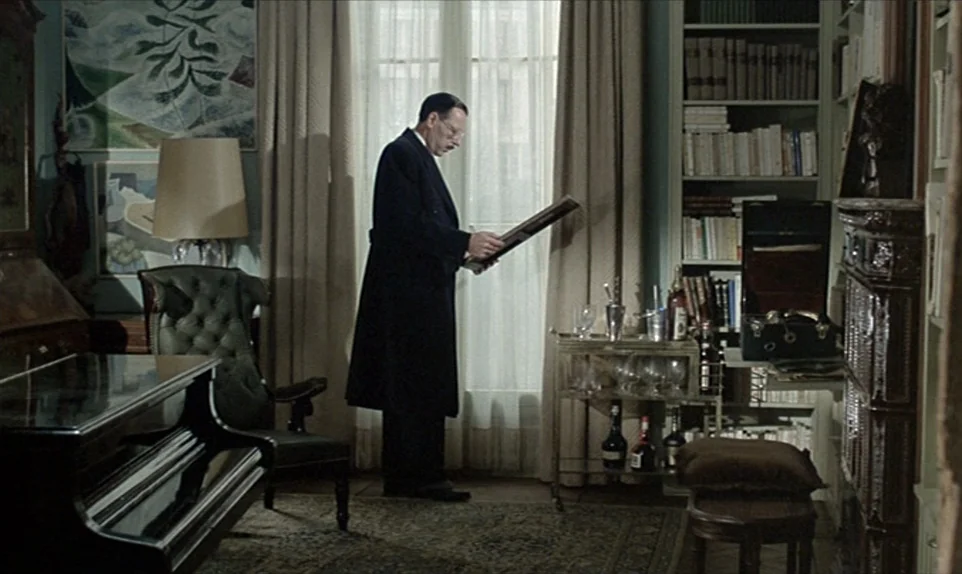 Mr. Klein (1976) movie still: medium — A study filled with bookcases, a piano, and a bar cart near a window. A middle-aged man stands in profile…