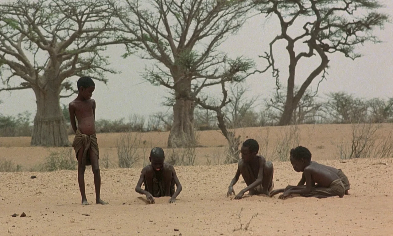 Coup De Torchon (1981) movie still: wide — A dry, arid landscape features sparse vegetation and baobab trees under a gray sky. Four children with…