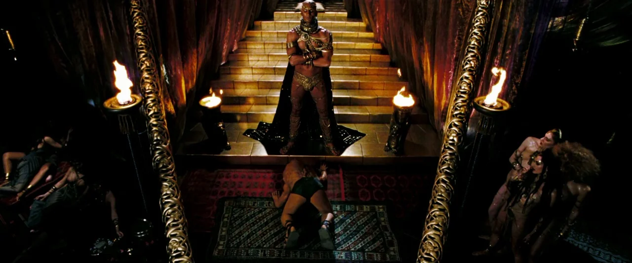 300 (2006) movie still: wide — A dark, opulent fantasy chamber features a stepped dais framed by tall, ornate gold pillars. A man stands proudly…