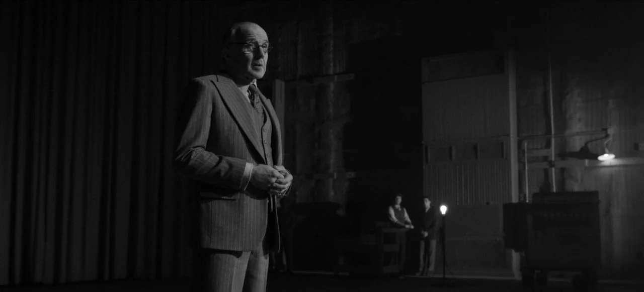 Mank (2020) movie still: medium — A dark, expansive film set features industrial architecture and heavy fabric curtains. An elderly man in a…