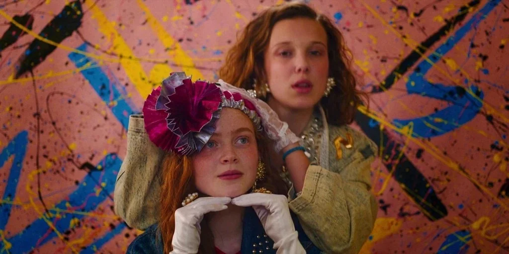 Stranger Things (2019) movie still: medium — The background consists of a wall covered in pink, blue, and yellow abstract paint. A teenager with red…