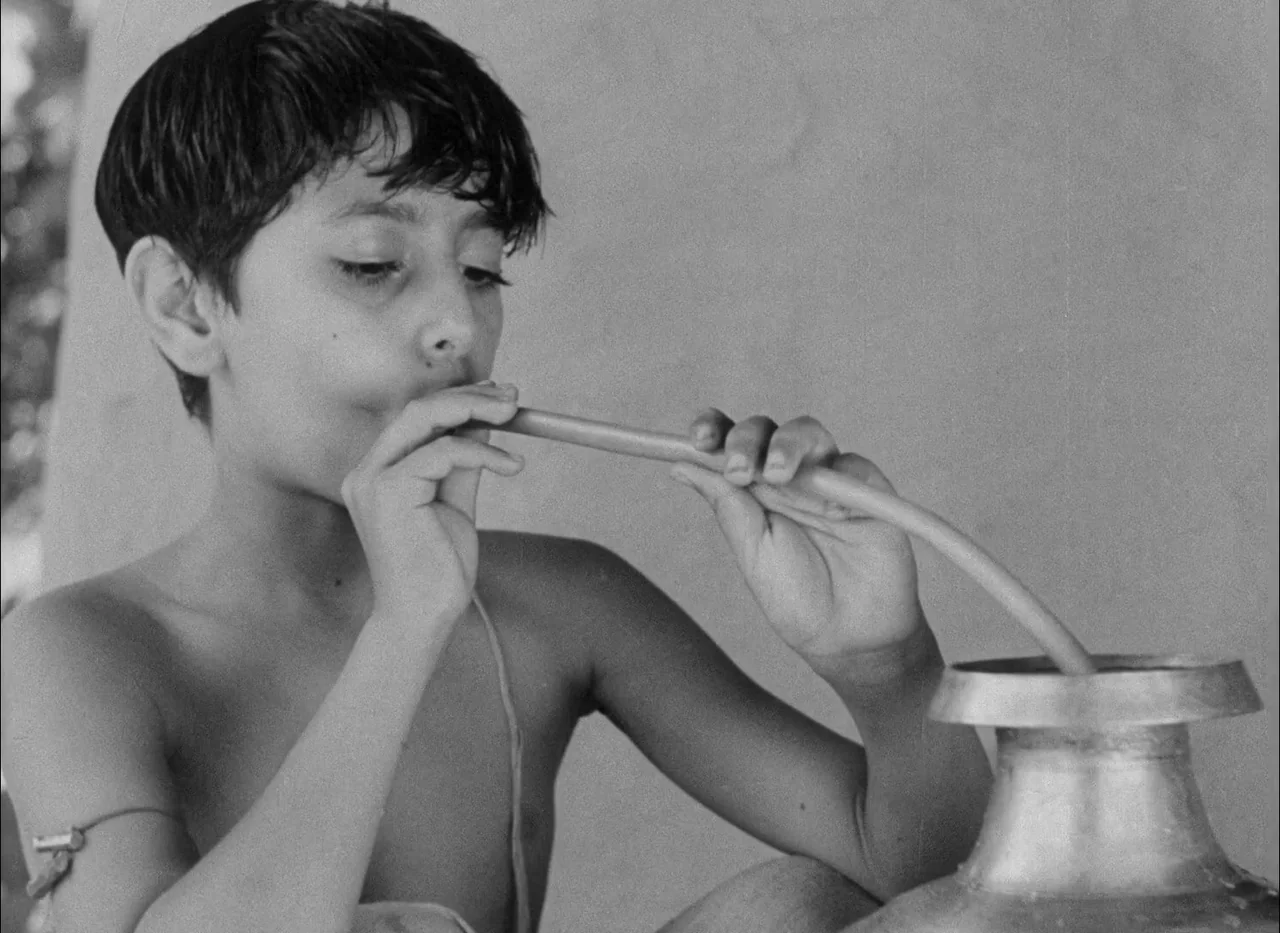 Aparajito (1956) movie still: closeup — An outdoor structure with textured walls serves as the backdrop for a young boy. The boy holds a rubber tube…