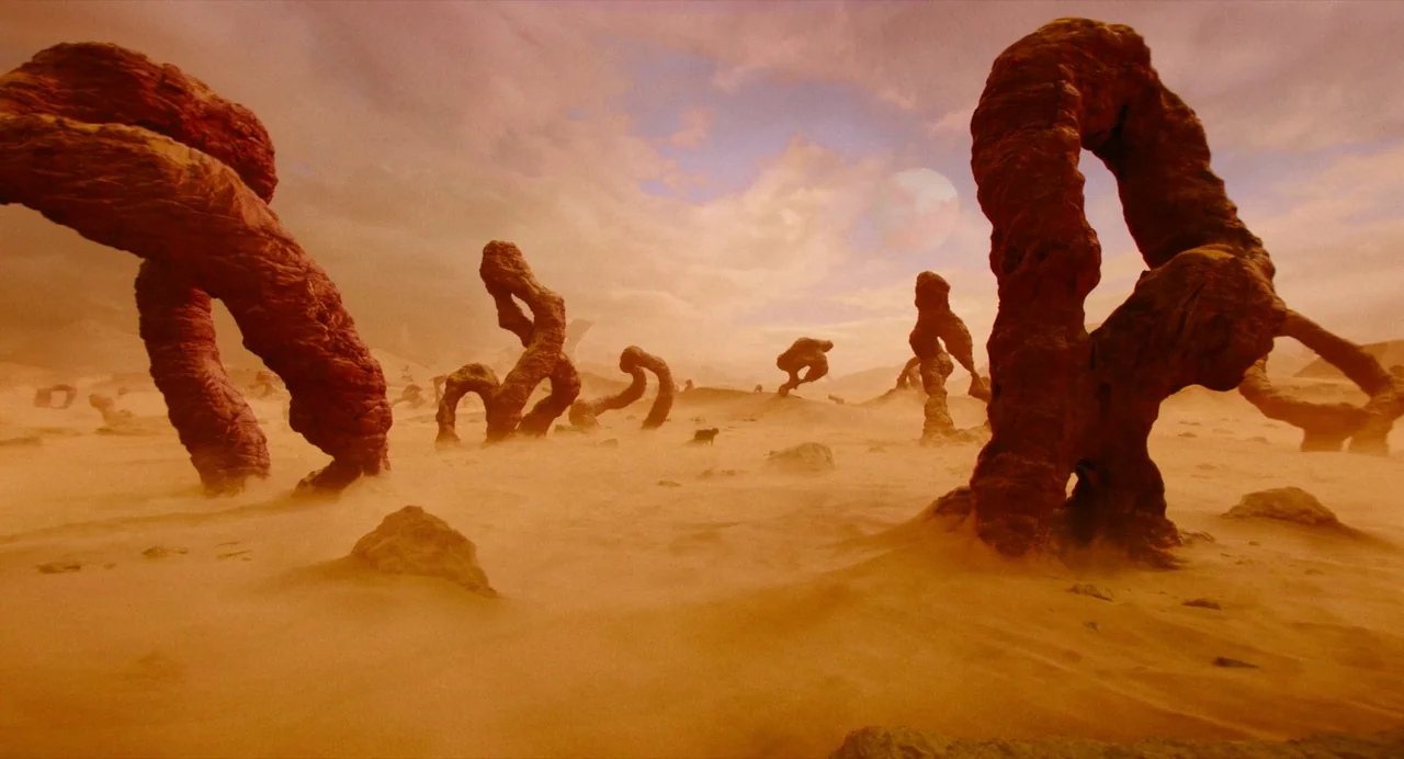 Beetlejuice Beetlejuice (2024) movie still: wide — An arid, sandy desert landscape features large, twisted reddish rock formations scattered…