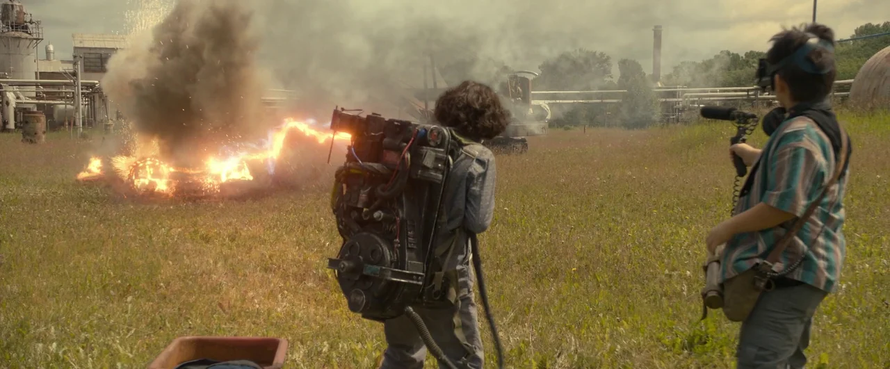 Ghostbusters: Afterlife (2021) movie still: fullbody — An overgrown field in front of an industrial facility is filled with hazy smoke. One teenager…