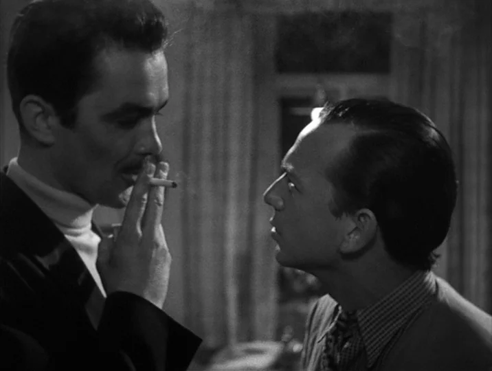 To Joy (1950) movie still: medium — A dimly lit room with draped curtains visible in the blurred background. Two men stand in close proximity as one…