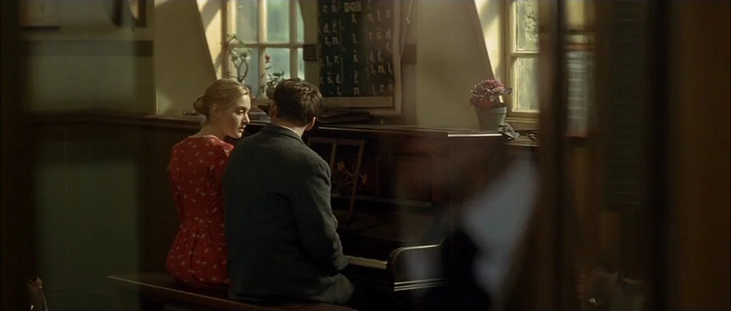 Jude (1996) movie still: medium — A wood-paneled room features a piano positioned beneath two large, multi-paned windows. A man and a woman sit…