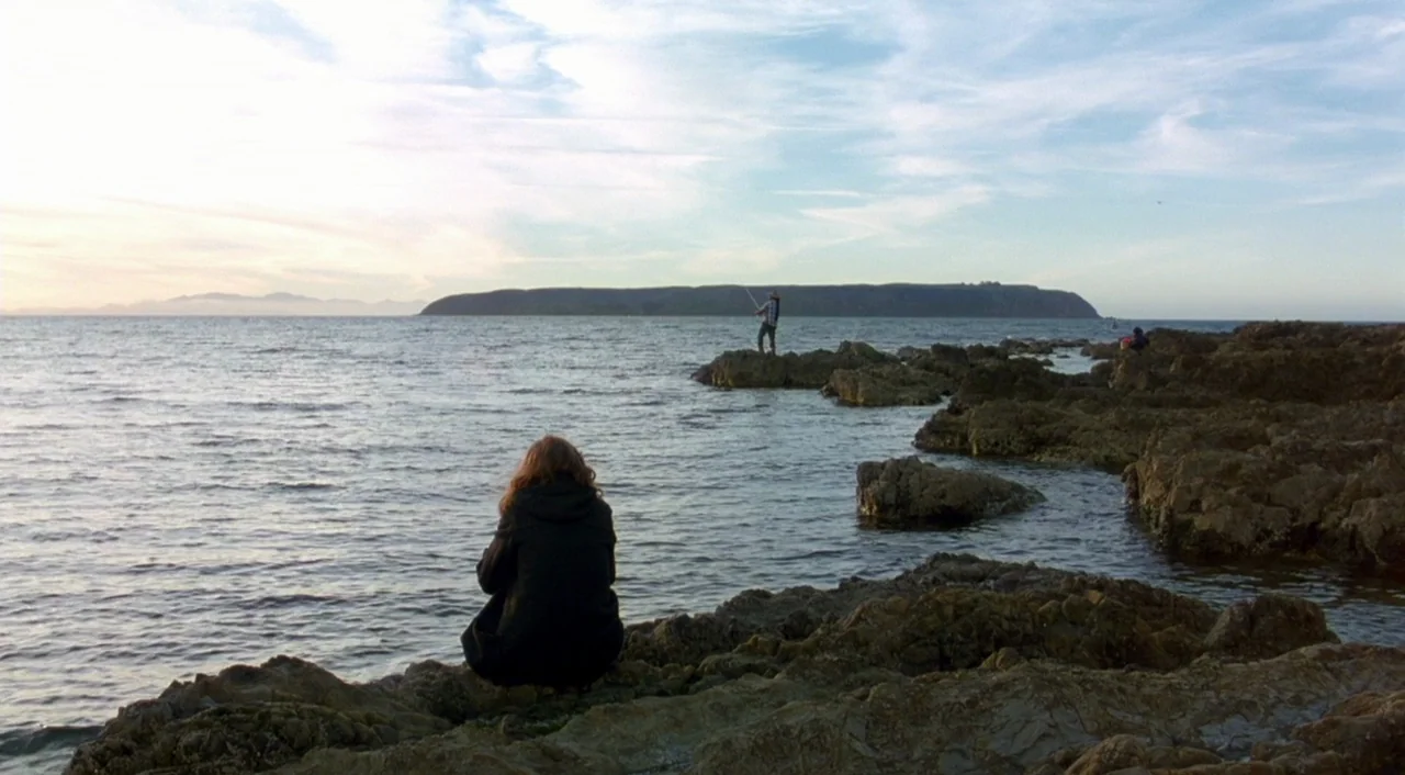 Eagle Vs. Shark movie still: wide — A rocky shoreline meets the open ocean under a partially cloudy sky. A person sits on the rocks while another…