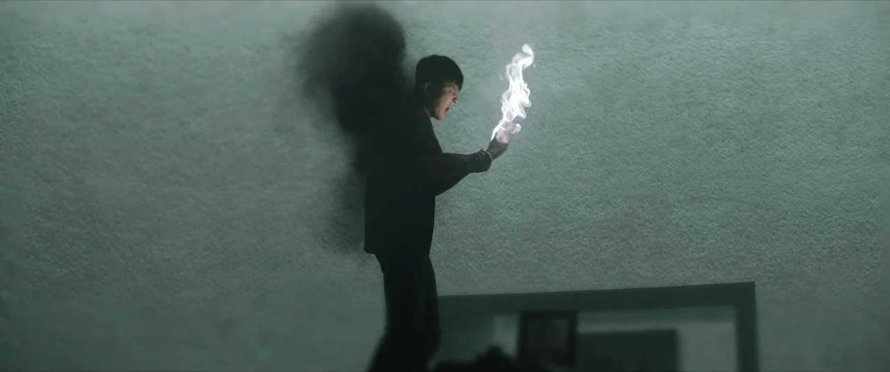 The Divine Fury (2019) movie still: fullbody — A man stands in an abstract, dimly lit space with a muted textured wall. The subject holds a bright…