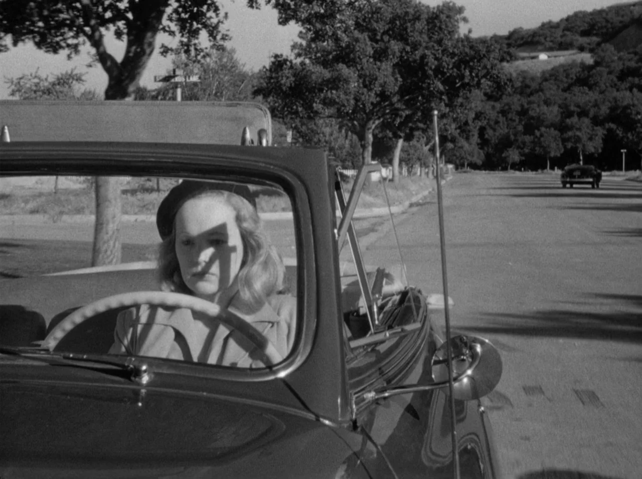 Gun Crazy (1950) movie still: medium — A woman drives an open-top convertible along a quiet tree-lined road during the day. She holds the steering…