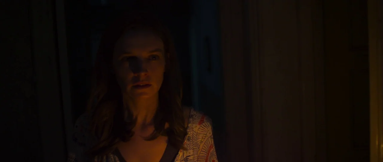 A Dark Song (2016) movie still: medium — A dark, residential hallway with very little light visible beyond the main subject. The woman stands in the…