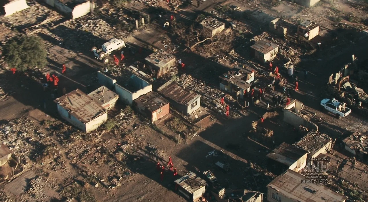 District 9 (2009) movie still: establishing — A dense cluster of small corrugated metal shacks sits on uneven dirt ground. Multiple individuals…