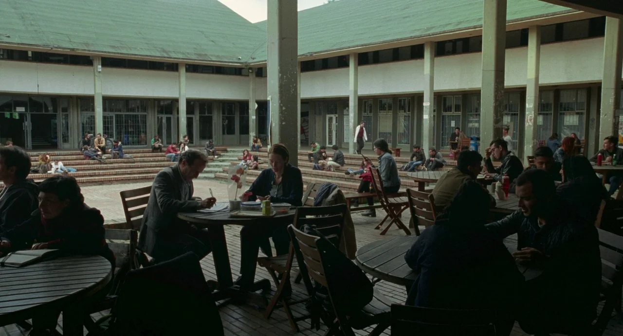 Memoria (2021) movie still: wide — An outdoor university courtyard features tiered stone seating and circular wooden tables. Many students sit at…