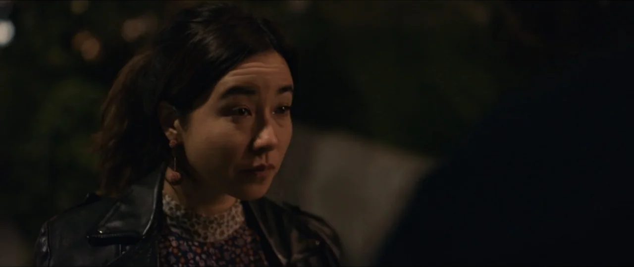 Plus One (2019) movie still: closeup — An outdoor night setting featuring a woman in a leather jacket on a sidewalk. The woman stands facing an…