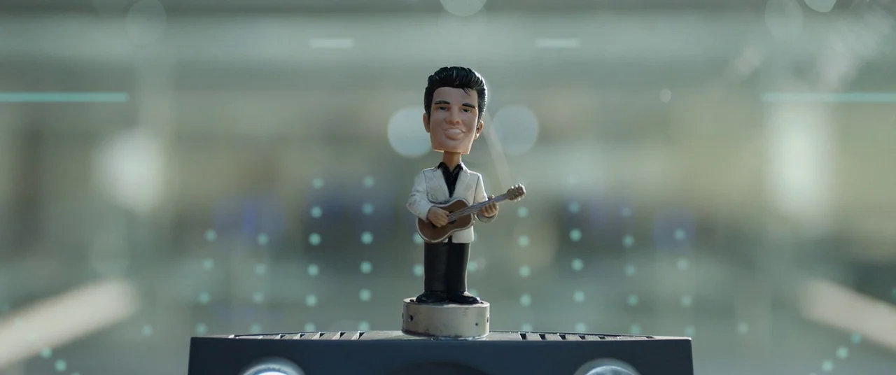 Oblivion (2013) movie still: closeup — A miniature Elvis bobblehead stands atop a black device inside a glass display. The figure faces forward…