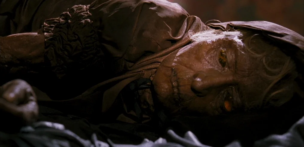 Tideland (2005) movie still: closeup — A dark space features a wrinkled individual lying down on a dark fabric surface. The person has leathery skin…