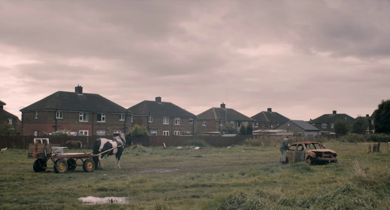 The Selfish Giant (2013) movie still: wide — A muddy field sits before a row of brick council houses under a cloudy sky. A man inspects the wreckage…