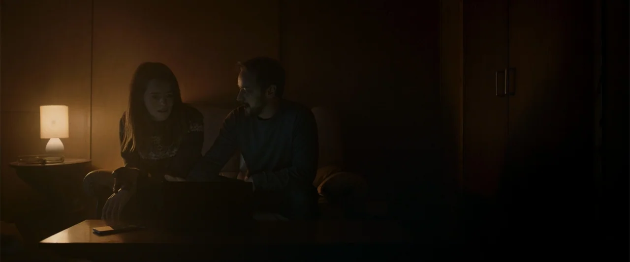 My Son (2021) movie still: medium — A dimly lit room contains a sofa and a wooden table lamp at night. A young couple sits together on the couch and…