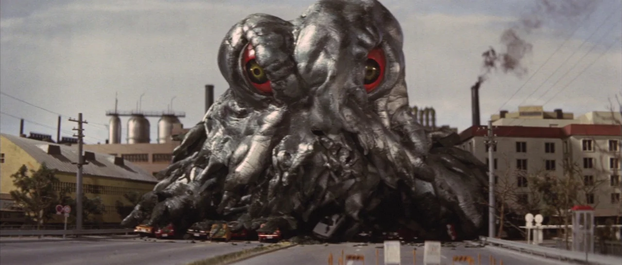 Godzilla Vs Hedorah (1971) movie still: fullbody — A massive, viscous metallic monster looms over an urban road near industrial smokestacks. The…