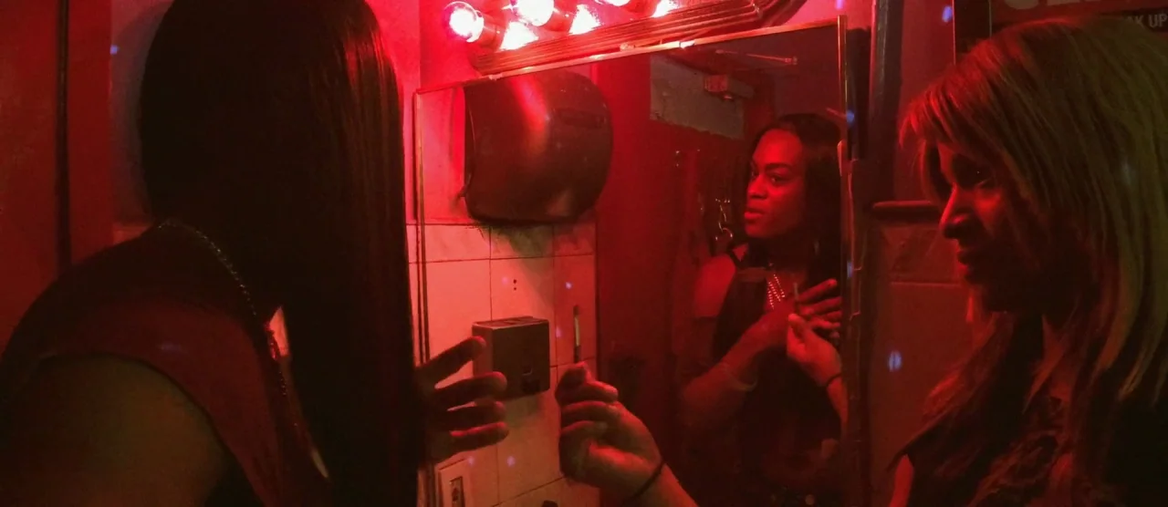Tangerine (2015) movie still: medium — A dimly lit restroom features a mirror reflecting two people under harsh red lights. The subjects stand near…