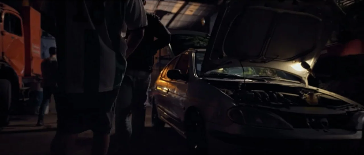 Gunman (2025) movie still: medium — A dimly lit garage with a sedan parked and its hood propped open. Two men stand beside the vehicle while a work…