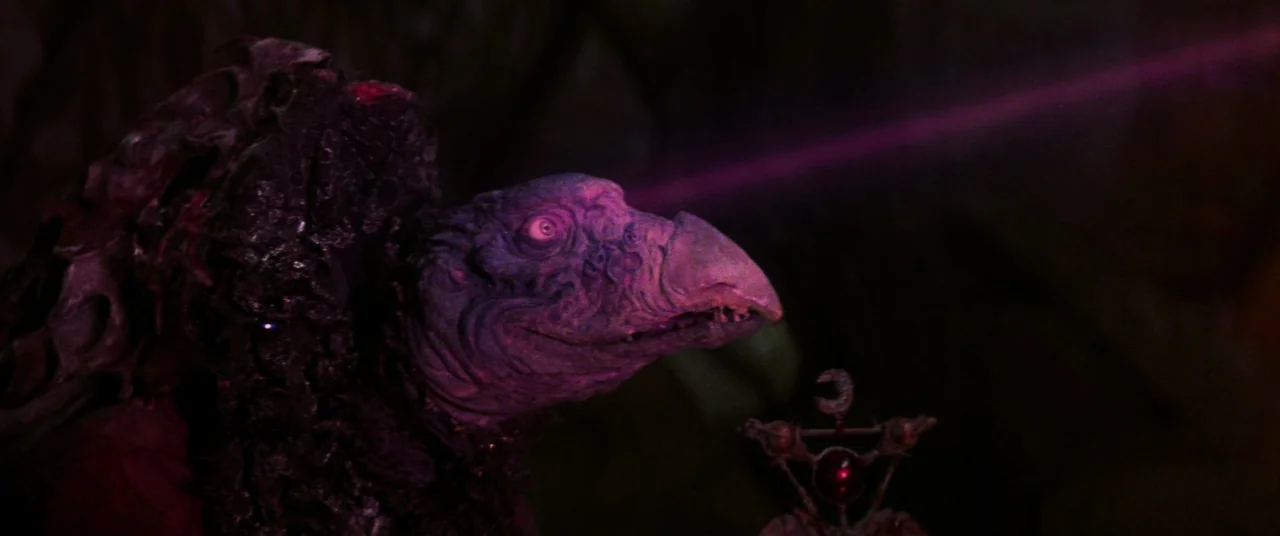 The Dark Crystal (1982) movie still: closeup — A dimly lit, dark space houses a large, textured avian puppet creature. The creature turns its head…