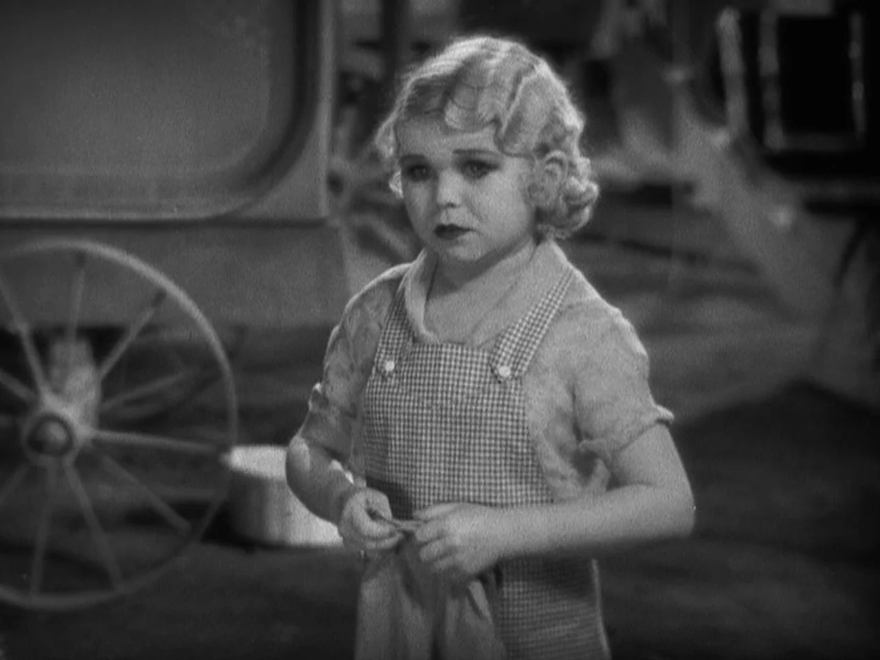 Freaks (1932) movie still: medium — A child stands in a sparse outdoor area with a wooden wheel in background. The child wears a checkered apron and…