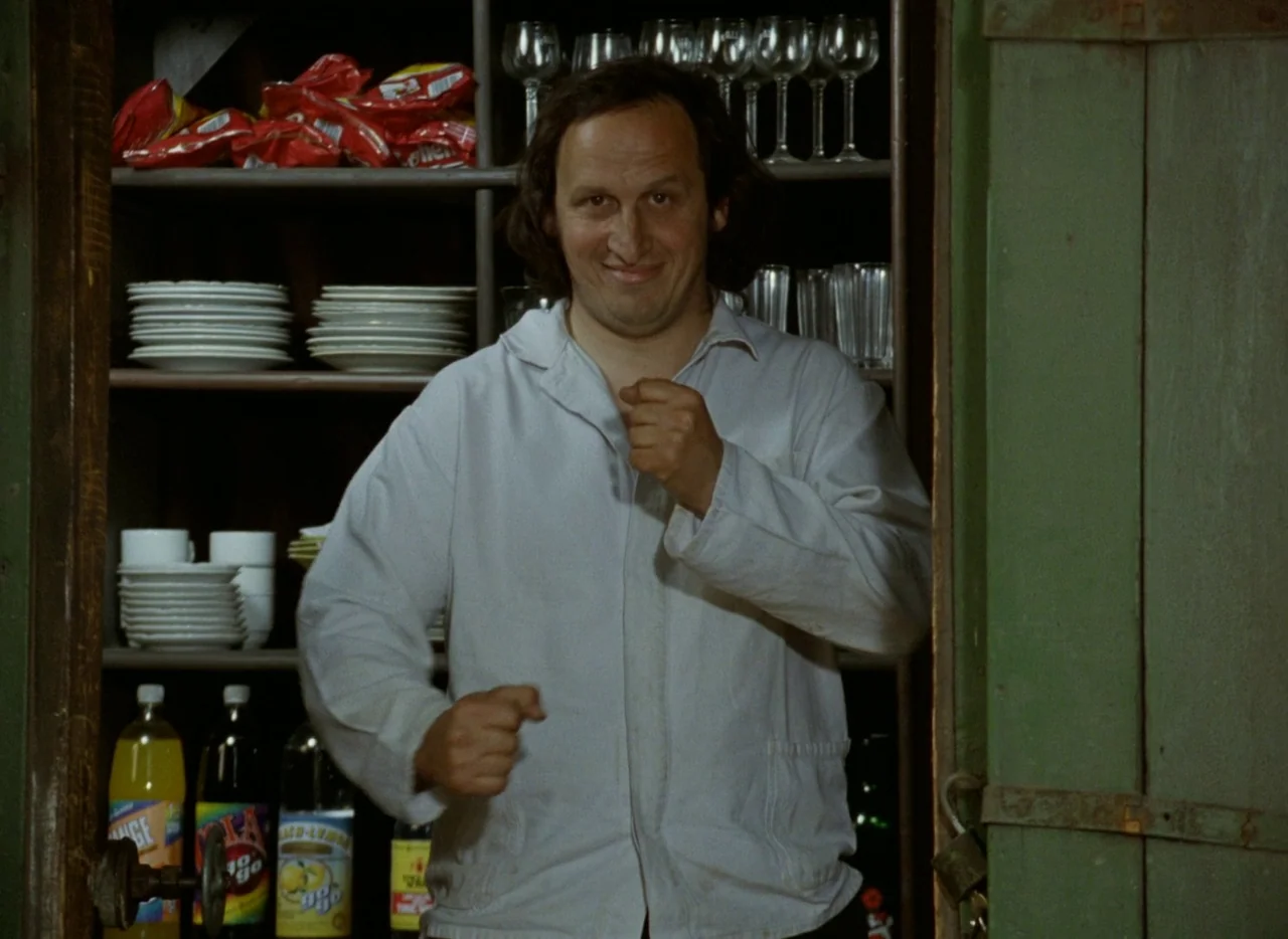 Faust (1994) movie still: medium — A man stands in front of a wooden shelving unit filled with plates and drinks. He holds his fists up to his chest…