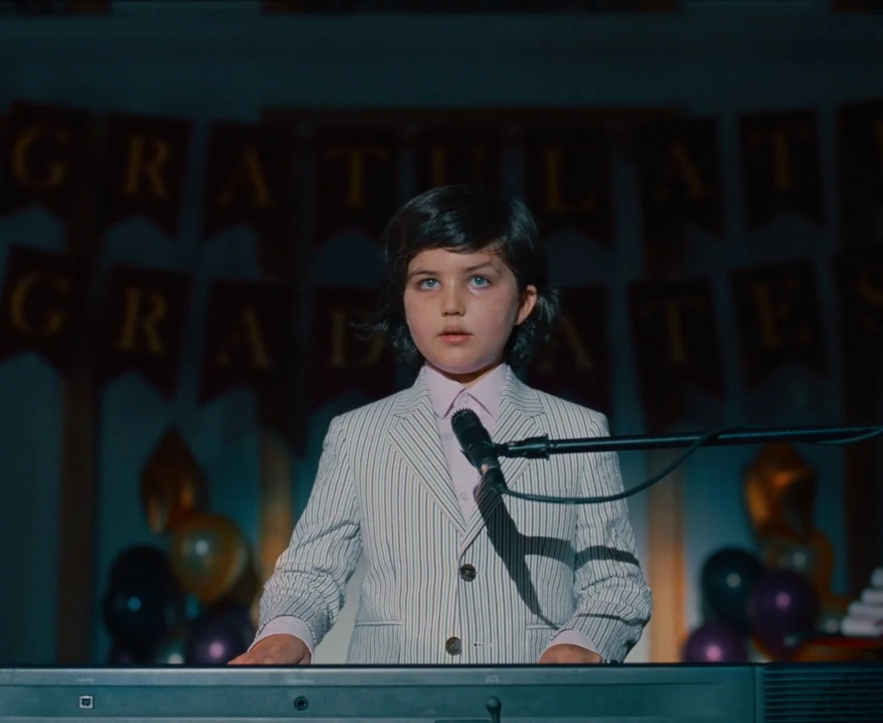 Monica (2022) movie still: medium — A dark event hall features a banner reading congratulations graduates hanging on the back wall. A young boy…