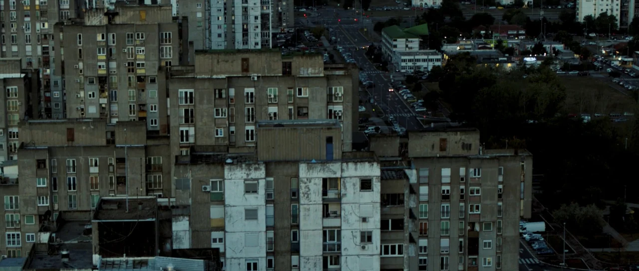 Hellraiser (2022) movie still: establishing — A dense cluster of weathered concrete brutalist apartment blocks fills the frame at dusk. A road with…