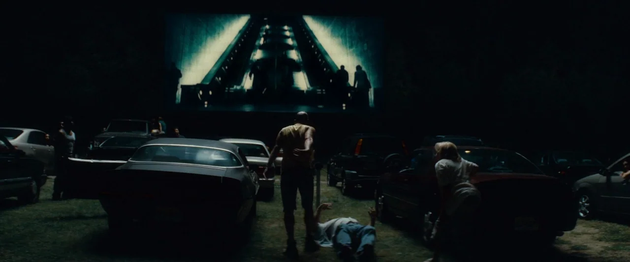 Out of the Furnace (2013) movie still: wide — A dark, grassy area at a drive-in theater with cars parked in rows. A man stands over a prone figure…