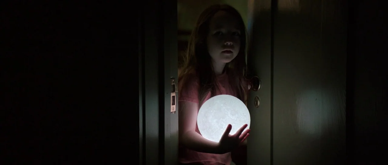 The Boogeyman (2023) movie still: medium — A young girl stands in a dark doorway frame. She holds a glowing circular moon lamp in front of her.