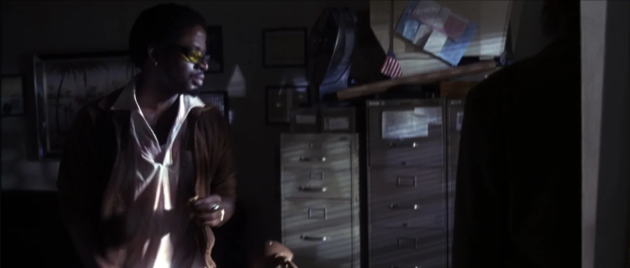 Gang Related (1997) movie still: medium — A cramped office space is cluttered with metal filing cabinets and an electric fan. A man wearing tinted…