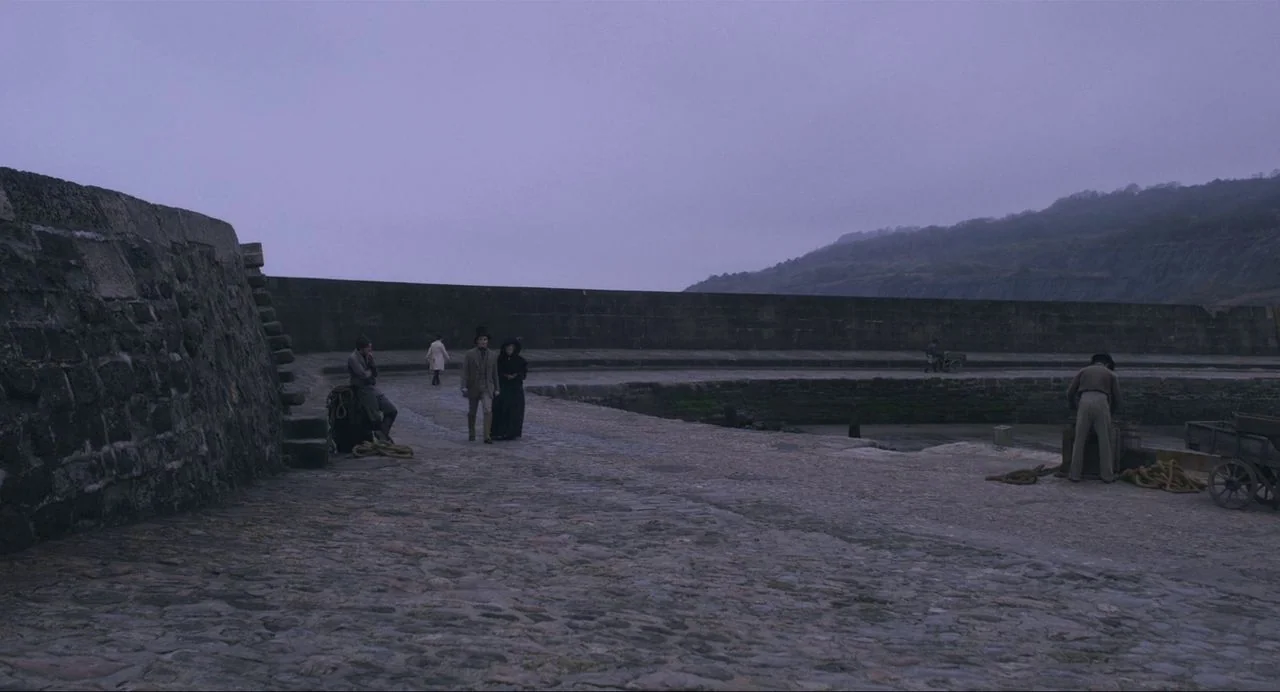 Ammonite (2020) movie still: wide — A desolate stone harbor pier extends under a flat, overcast sky near distant cliffs. A couple walks along the…
