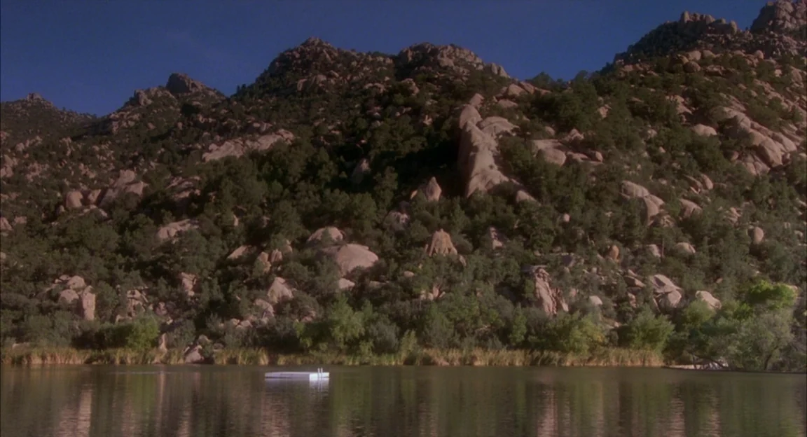 Creepshow 2 (1987) movie still: establishing — A calm lake sits beneath a rugged, rocky mountain slope covered with scattered trees. A small white…