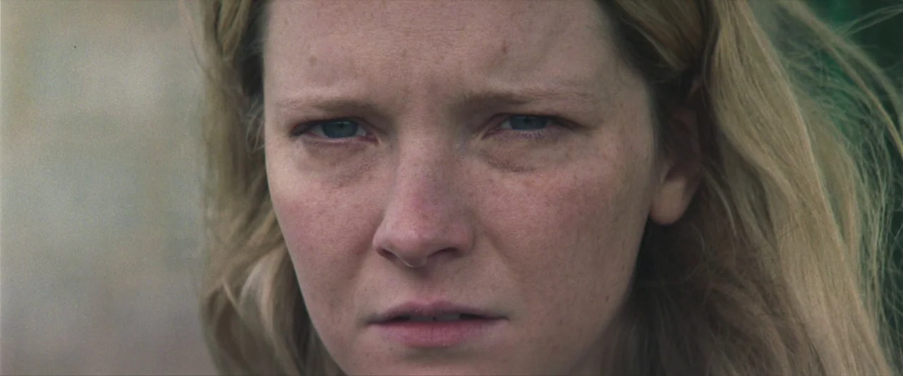 Starve Acre (2023) movie still: closeup — A close-up view of a woman's face against a blurred green outdoor backdrop. She stares ahead while her…