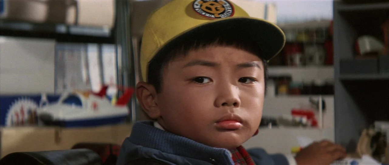 All Monsters Attack (1969) movie still: closeup — A child sits inside a room filled with various hobbyist items and toy models. The boy turns his…