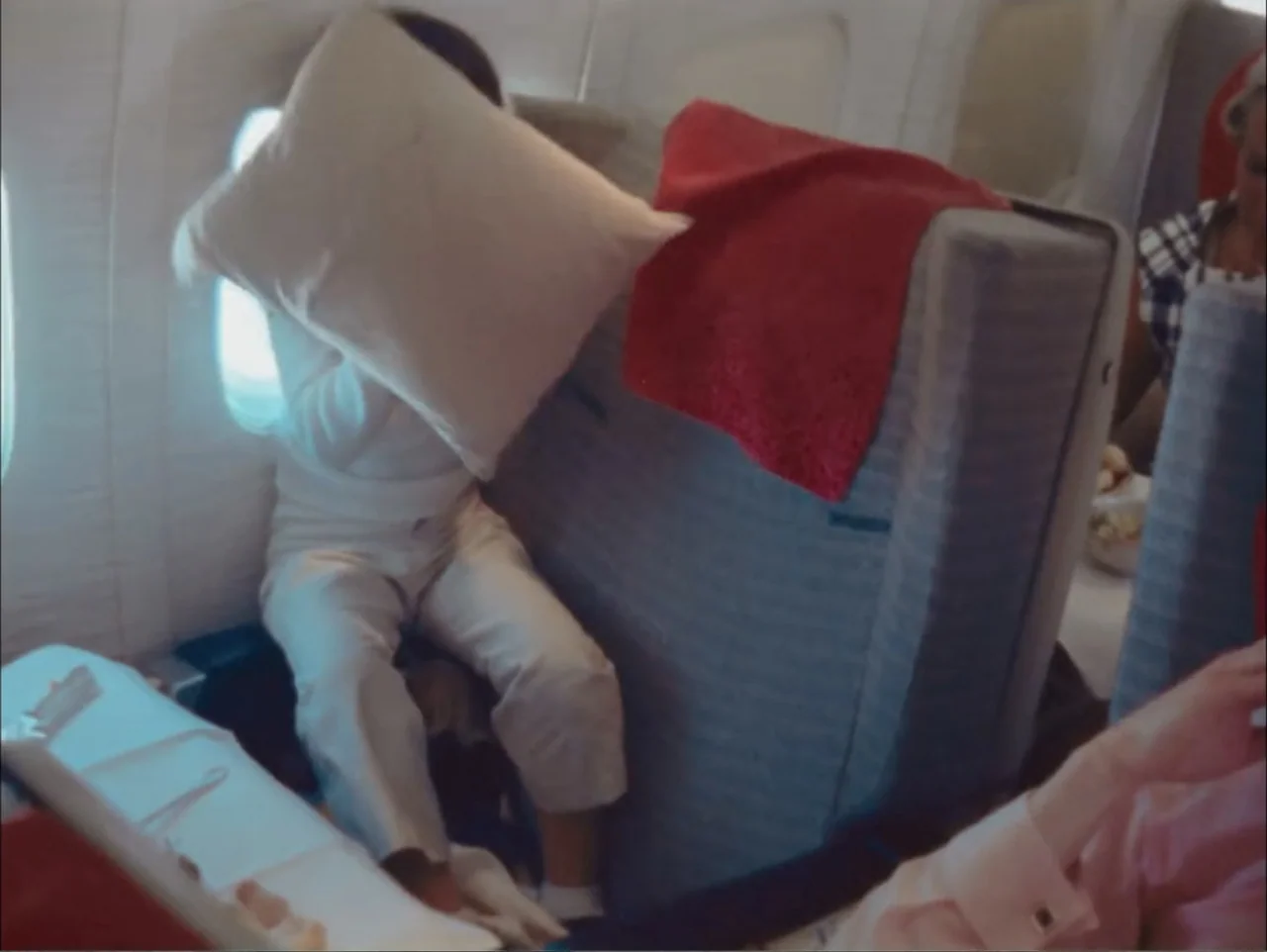 One to One: John & Yoko (2025) movie still: medium — An interior of an airplane cabin from the 1970s with grey fabric seating. A person sits in a…