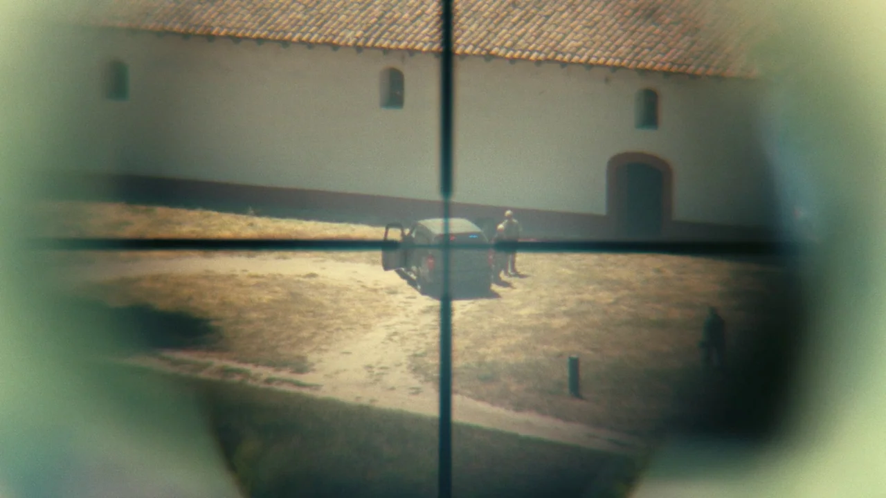 One Battle After Another (2025) movie still: wide — A stucco building with a tiled roof sits behind an open dirt courtyard. A person stands near an…