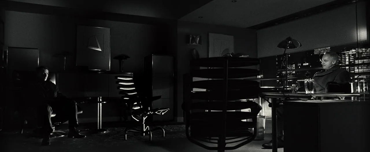 Casino Royale (2006) movie still: wide — A dark, modern workspace features a minimalist desk and chairs with city lights visible. Two men occupy the…