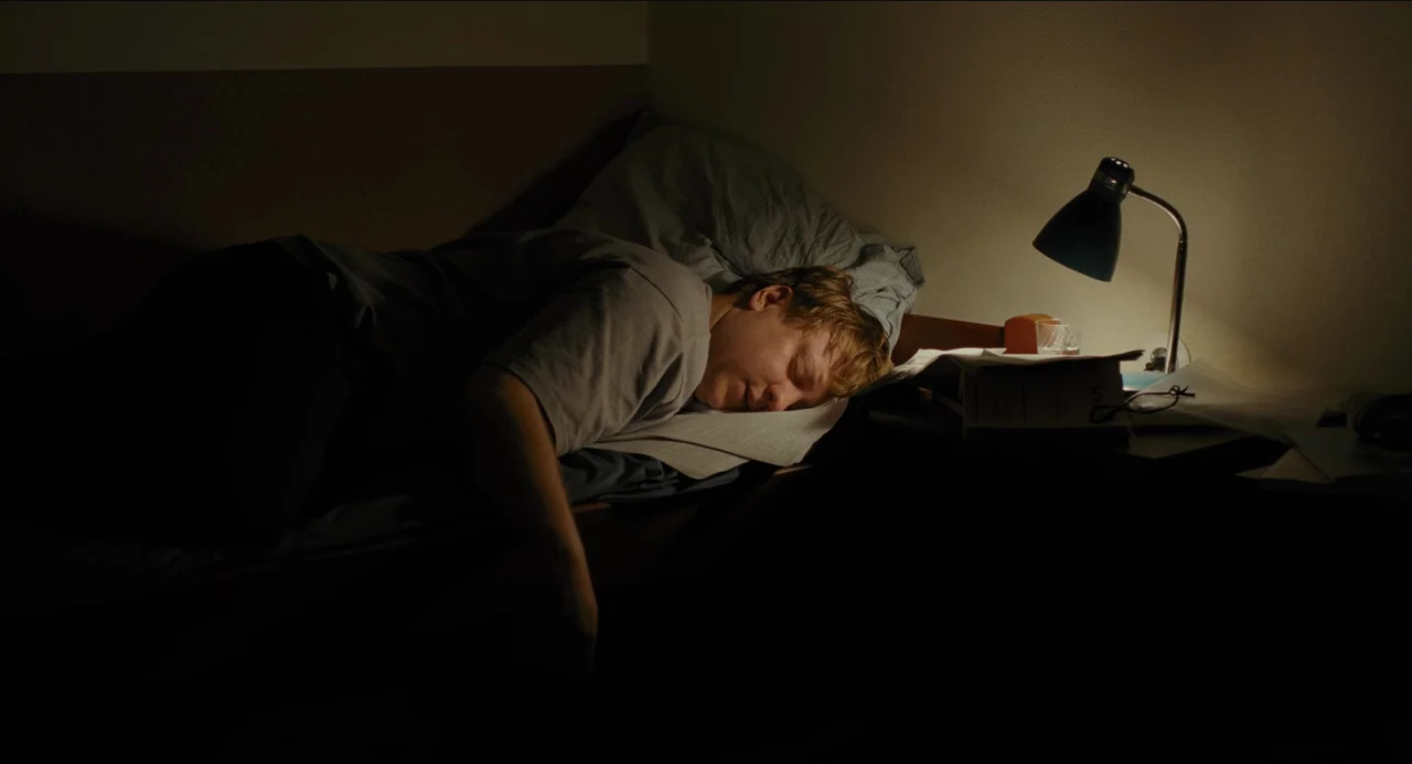 Afire (2023) movie still: medium — A man sleeps on a bed with his head resting on open books. A small lamp illuminates the surface of the paper near…