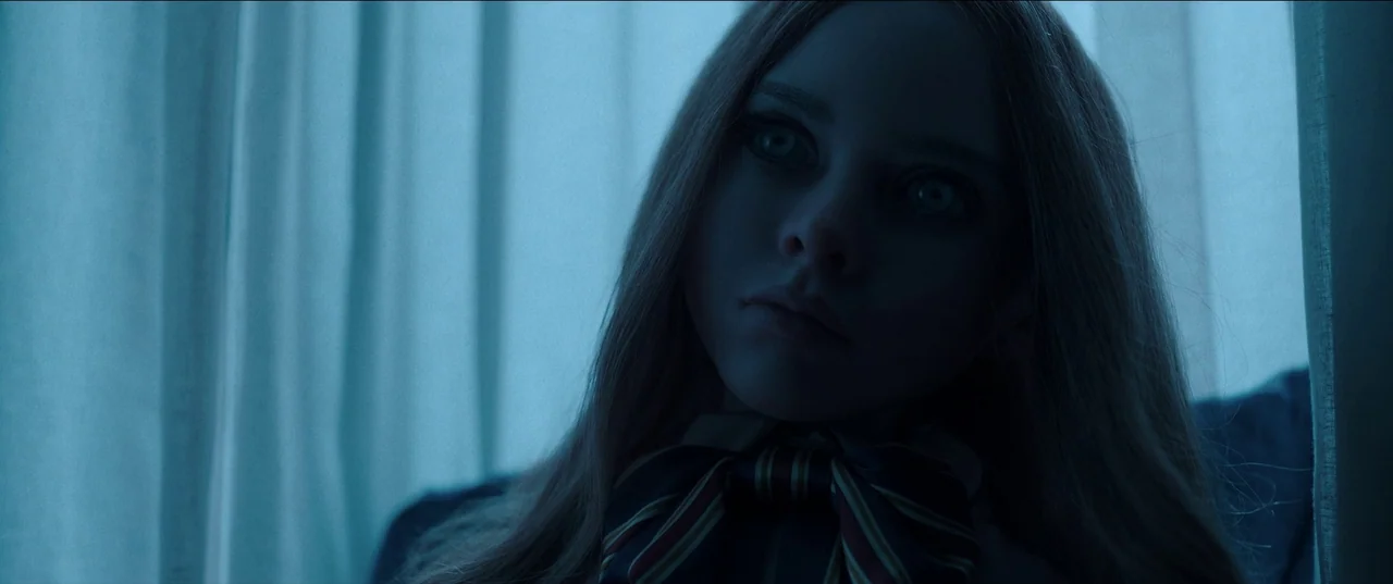 M3gan (2022) movie still: closeup — A close portrait of a doll-like figure stands before a dark fabric background. The subject stares ahead with…