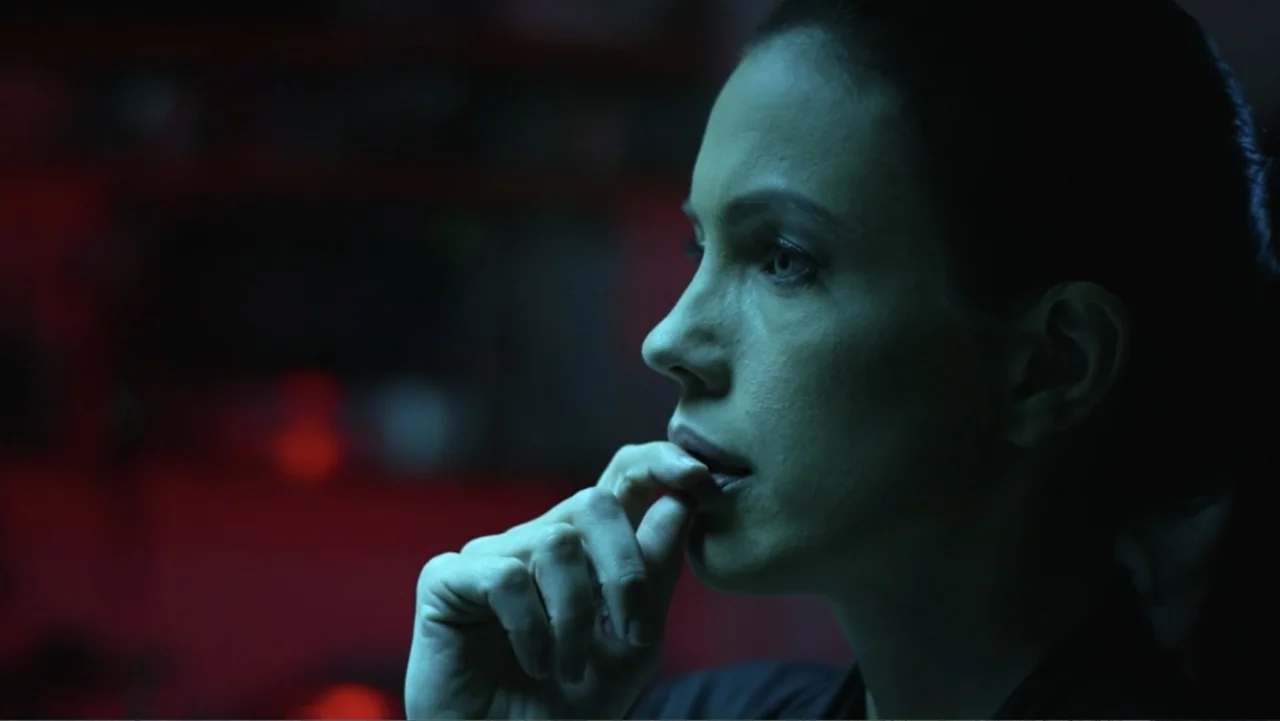 Entre las sombras (2025) movie still: closeup — A dimly lit interior space features a dark room with red shadows. A woman rests her fingers near her…