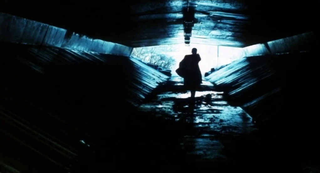 Narc (2002) movie still: wide — A dark concrete underpass with slanted walls leads toward a bright patch of daylight. A person walks through the…